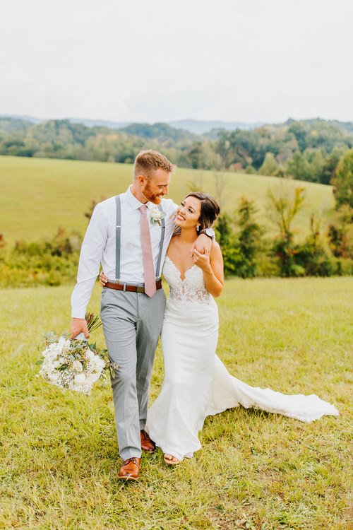 Couple in wedding attire walks through a grassy field with rolling green hills and trees in the background under an...