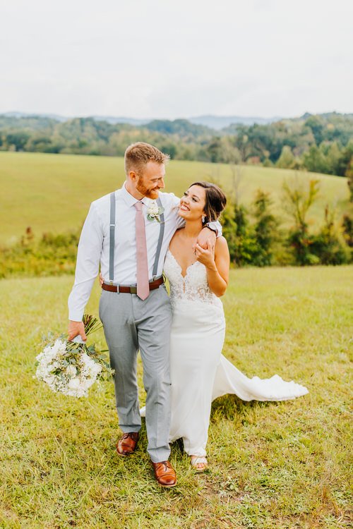 Wedding couple in a white dress and gray suspenders walk across a green field with rolling hills and trees in the background.