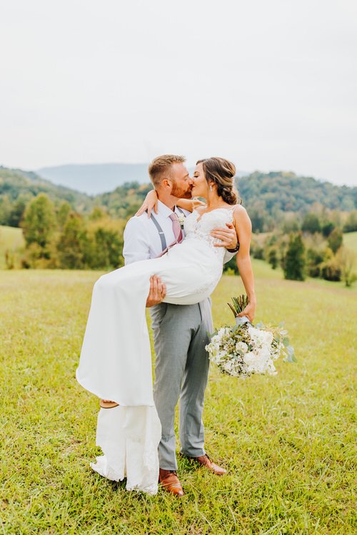 Groom carries bride in a grassy field with distant mountains and trees under an overcast sky. Bride holds a white floral...