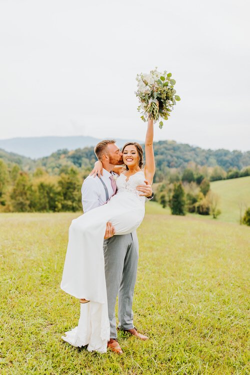 Groom in gray pants carrying bride in white gown, kissing her cheek. Bride holds bouquet up in a grassy field with hills.