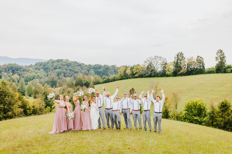 Wedding party on a grassy hill, arms raised. Bride, bridesmaids in pink, groomsmen in gray pants. Mountain backdrop.