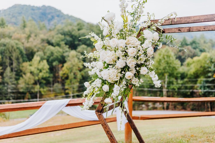 White floral arrangement on a rustic wooden wedding arch with green mountains and a wooden fence draped in white fabric...