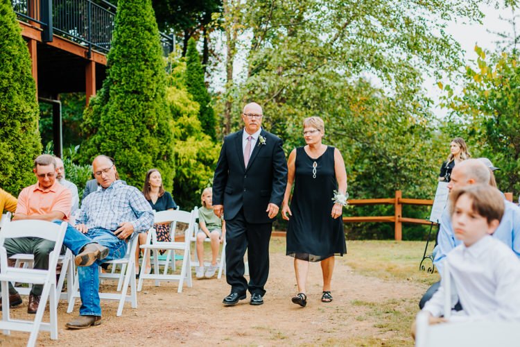 A man in a suit and a woman in a black dress walk down an outdoor aisle past seated guests and tall green trees. A wooden...