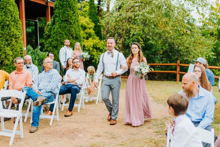 Wedding party member and bridesmaid walk an outdoor aisle at Moose Hollow Lodge, with seated guests and green trees.