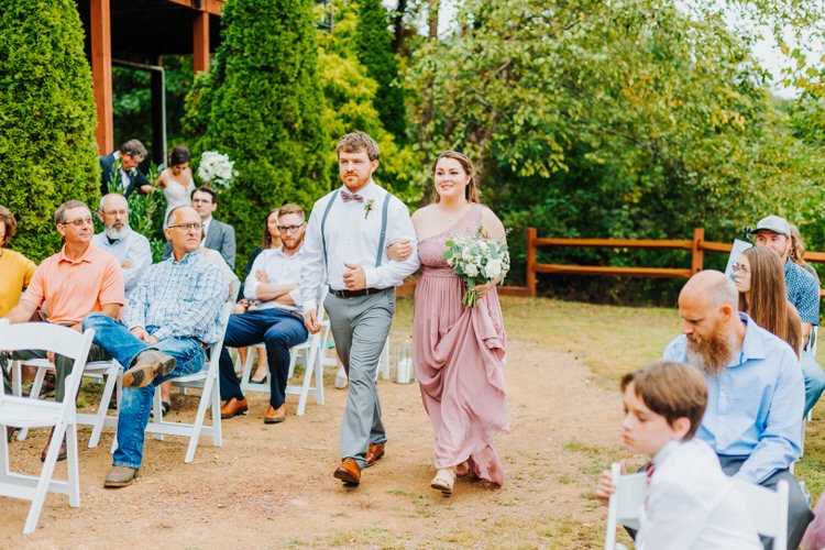 Man in suspenders and woman in pink dress with bouquet walk down outdoor aisle past guests at Moose Hollow Lodge.