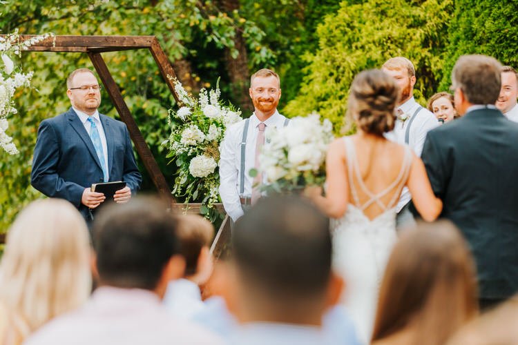 Outdoor wedding ceremony with bride walking towards groom, officiant, and guests. Wooden arch with white flowers.