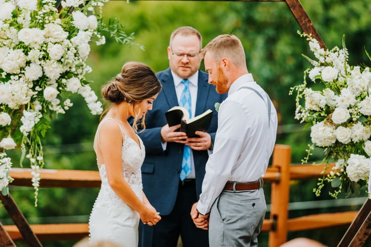 Bride in white dress, groom in white shirt, and officiant during an outdoor wedding ceremony under a floral arch.