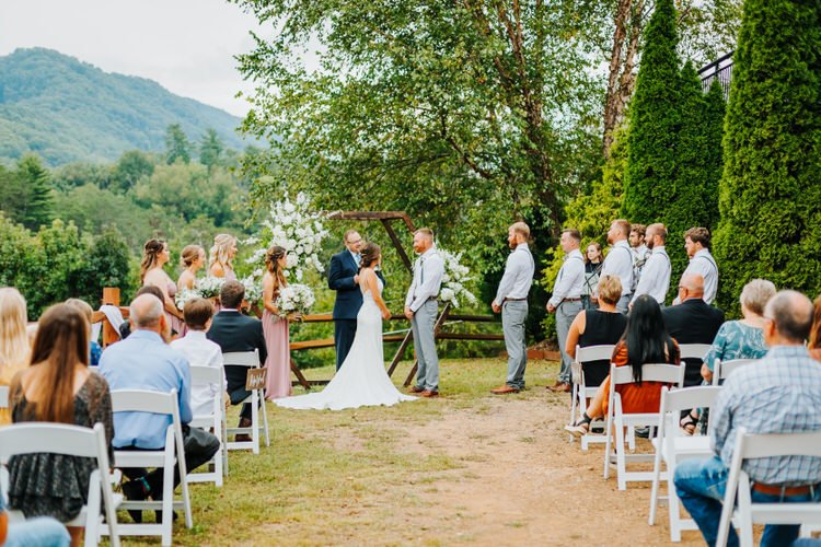 Outdoor wedding ceremony with bride, groom, wedding party, officiant, and guests. Wooden arch with white flowers,...