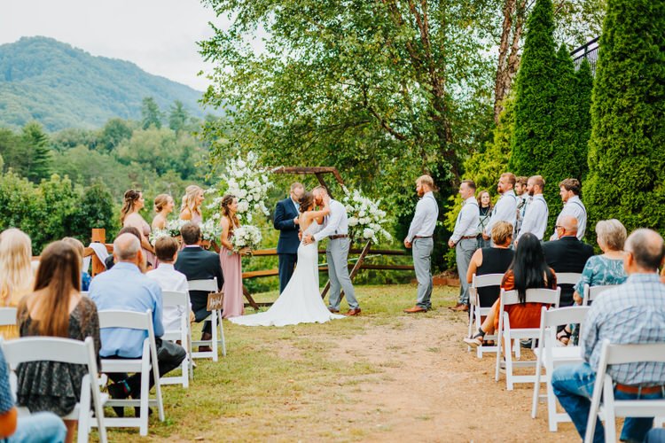 Outdoor wedding ceremony at Moose Hollow Lodge with bride and groom kissing, officiant, wedding party, and guests....