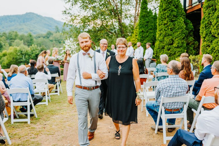 Groom with a woman walking down an outdoor wedding aisle with guests. Mountains and a cabin structure in background at...