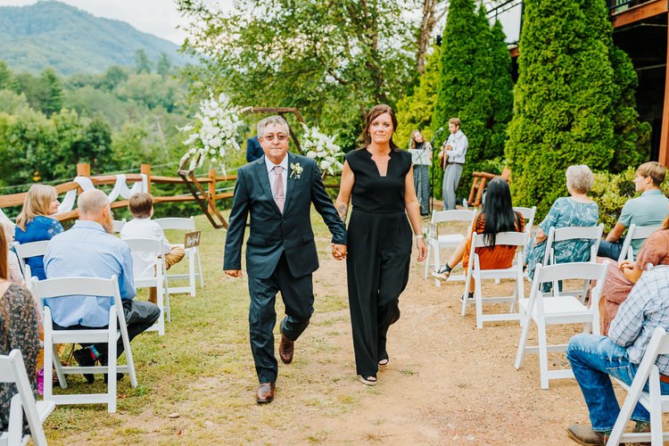 Man in suit and woman in black jumpsuit walk down outdoor aisle, guests seated, mountain backdrop at Moose Hollow Lodge.