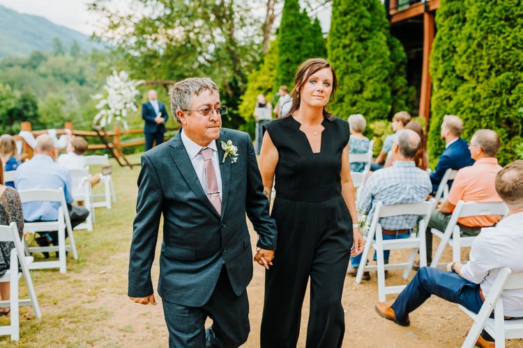 Man in suit and woman in black jumpsuit walk down outdoor aisle, guests seated, mountain view. Moose Hollow Lodge,...