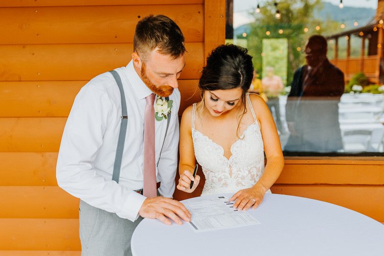 Couple signing papers at a white table, groom in suspenders, bride in white dress, against a log cabin wall at Moose...