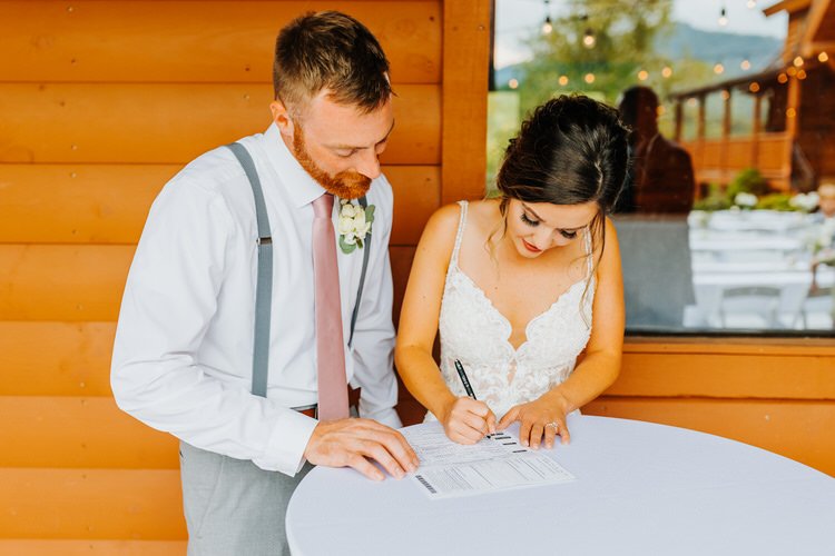 Bride and groom signing documents at a white table, next to a log cabin wall at Moose Hollow Lodge. Mountains and string...