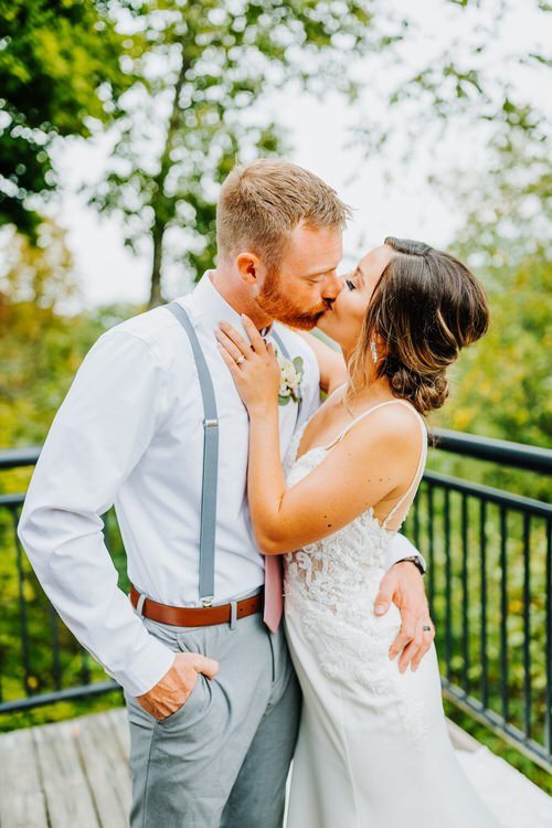 Man in suspenders and woman in wedding dress kiss on an outdoor deck with a black railing and green trees.