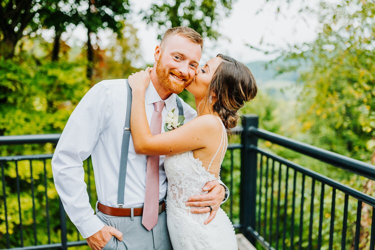 Couple in wedding attire on a Moose Hollow Lodge deck with green foliage and distant mountains. Groom in suspenders,...