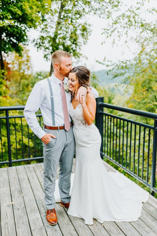 Groom kissing bride's forehead on a wooden deck with black railing, overlooking green trees and distant mountains. Moose...