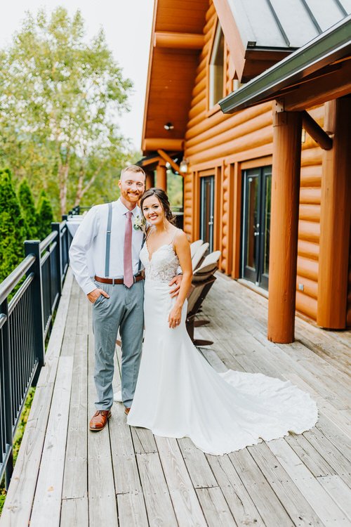 Bride and groom stand on a wooden deck in front of a log cabin, Moose Hollow Lodge, with green trees in background.