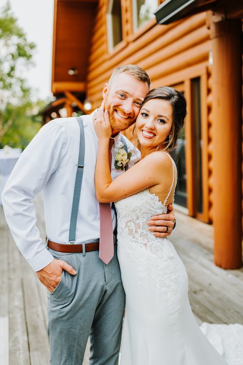 Smiling couple, a man in suspenders and woman in lace gown, embrace on a wooden deck in front of a log cabin at Moose...