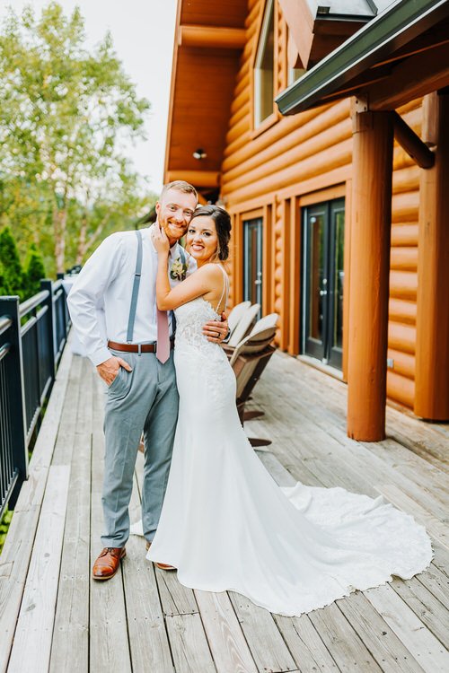 Couple in wedding attire on a wooden deck next to a large log cabin, likely Moose Hollow Lodge. Groom in suspenders.