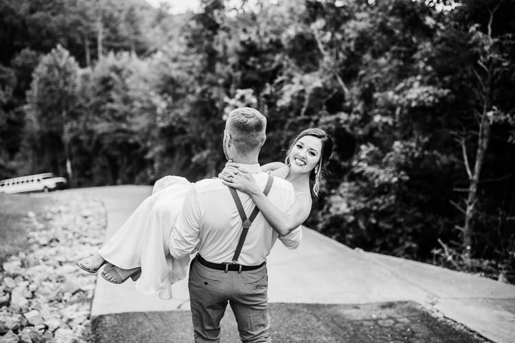 Groom carries bride down a paved path in a wooded area. A white limousine is visible in the background.