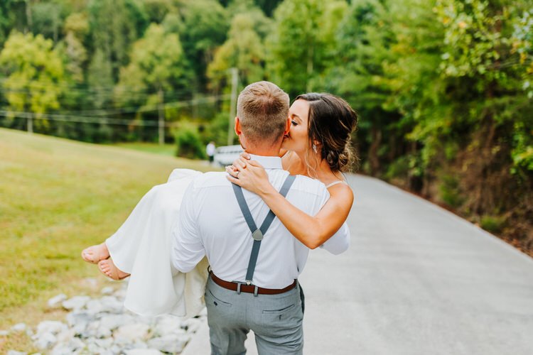 Man carrying a woman in a white dress on a paved road with green trees and grass. She leans in to kiss his ear.