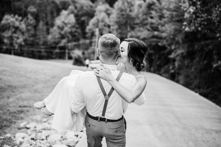 Black and white photo of a groom carrying his bride on a paved road bordered by grass and trees in the background.