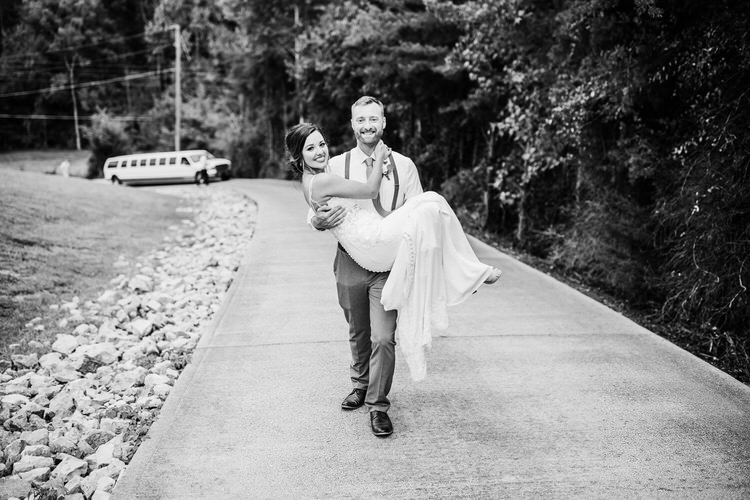 Black and white photo of a groom carrying his bride on a paved road lined with trees. A white limousine is in the background.