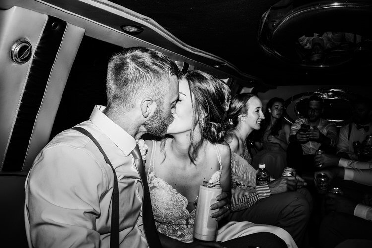 A black and white photo of a couple kissing in a limousine. The woman wears a wedding dress, the man a shirt and tie.