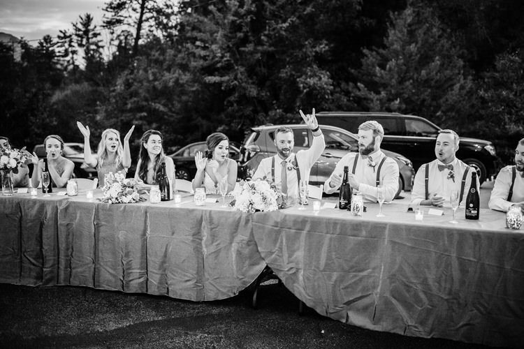Wedding party at an outdoor reception table with floral decor and candles. Guests make gestures, trees and cars in...