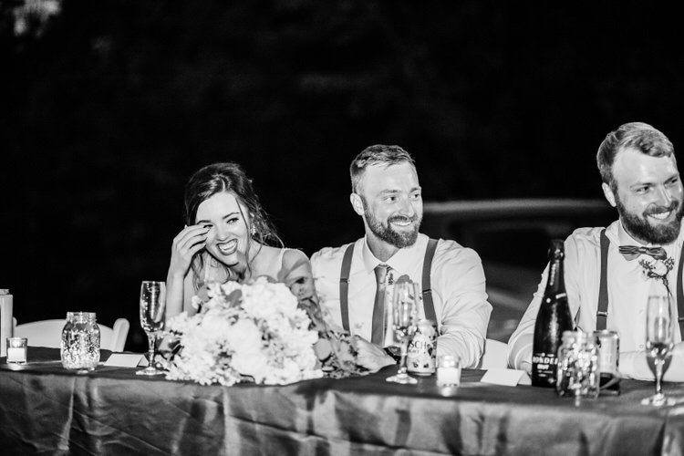 Bride and two men laughing at a reception table with floral centerpiece and drinks in a black and white photo at night.