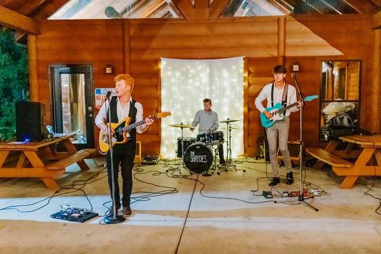 A live band, 'The Shaydes', performs on a concrete patio under a wooden lodge structure at Moose Hollow Lodge. String...