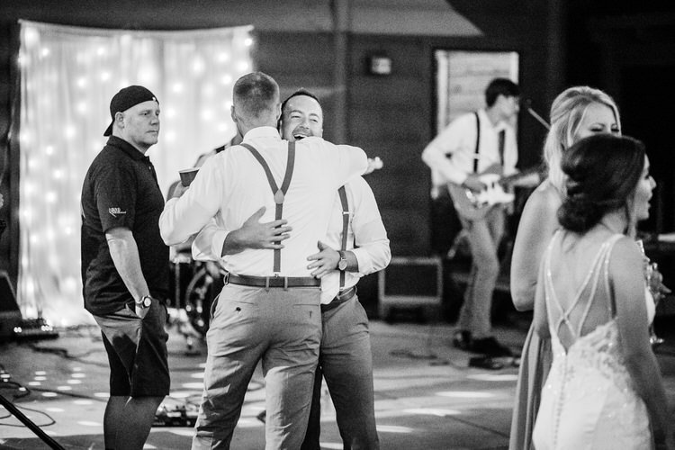 Guests hugging on a dance floor at a wedding reception. A band and string lights are in the background.