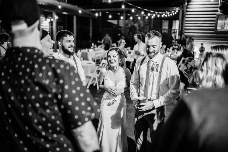 Bride in white dress, groom in suspenders, and guests conversing under string lights at a night outdoor reception at...