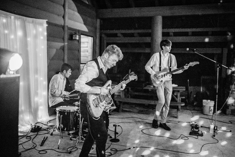 Live band with two guitarists and drummer performing at a log cabin venue. String lights on backdrop, spotlight on stage....